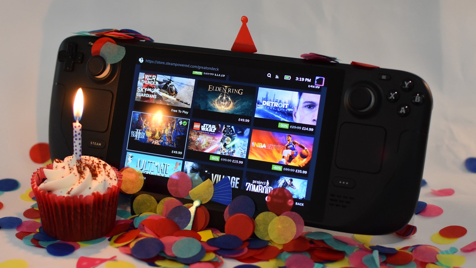 A Steam Deck surrounded by confetti and a cupcake with a lit birthday candle.