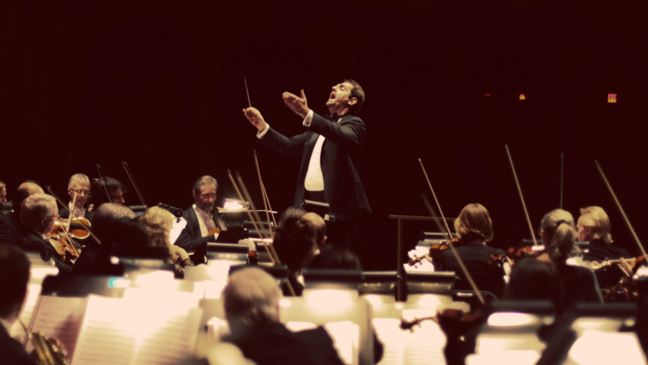 Conductor Andy Brick raises his hands in front of an orchestra at a Game ON concert