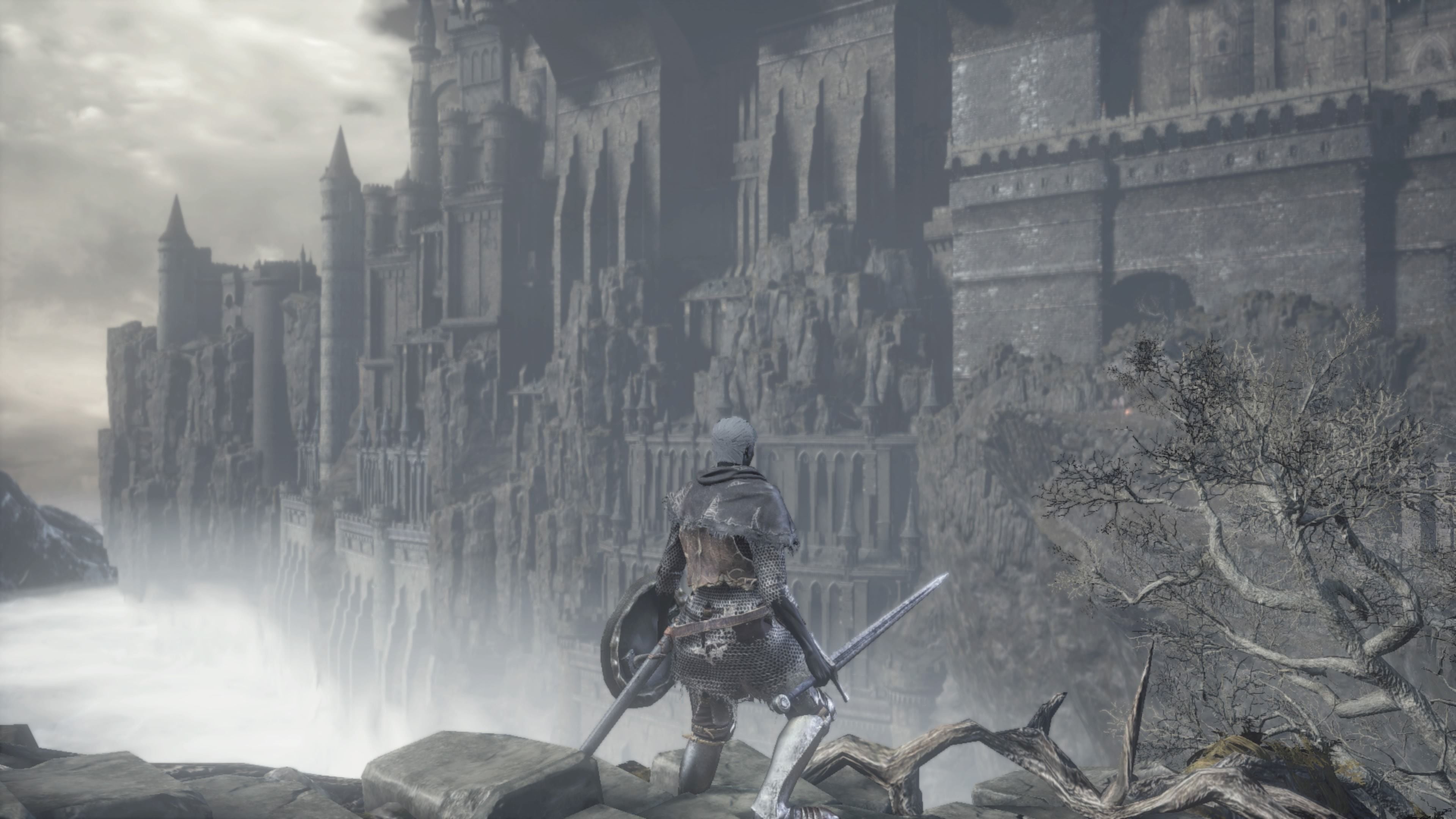 Um personagem em Dark Souls em armadura cinza ao lado de uma árvore cinza olhando para um penhasco cinza com um castelo cinza nele