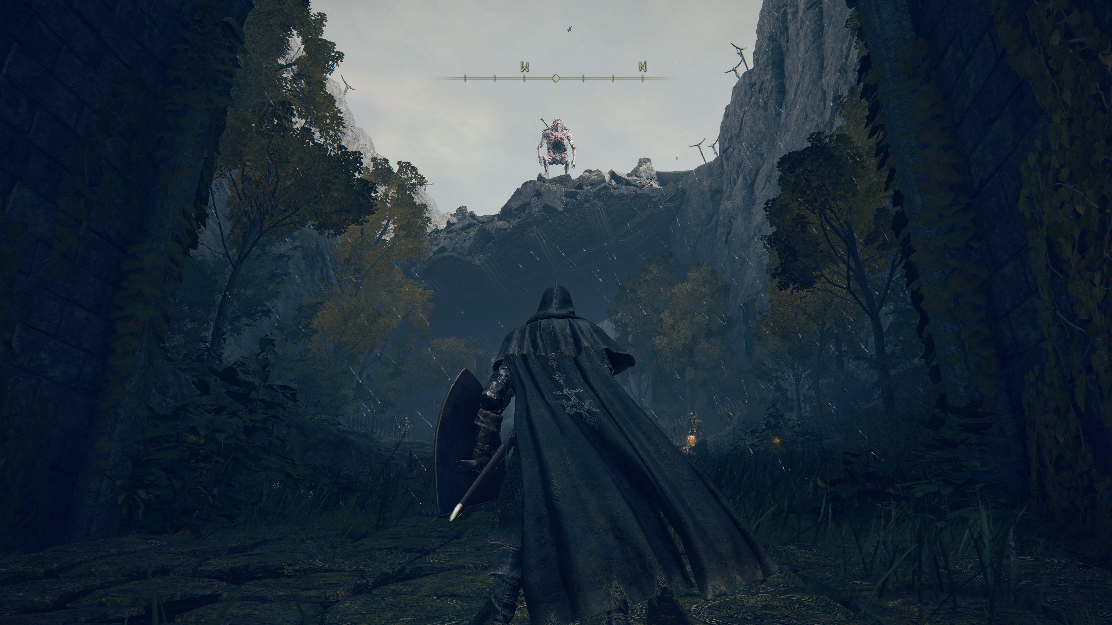 A cloaked warrior looks up at a large monster on top of a cliff in Elden Ring