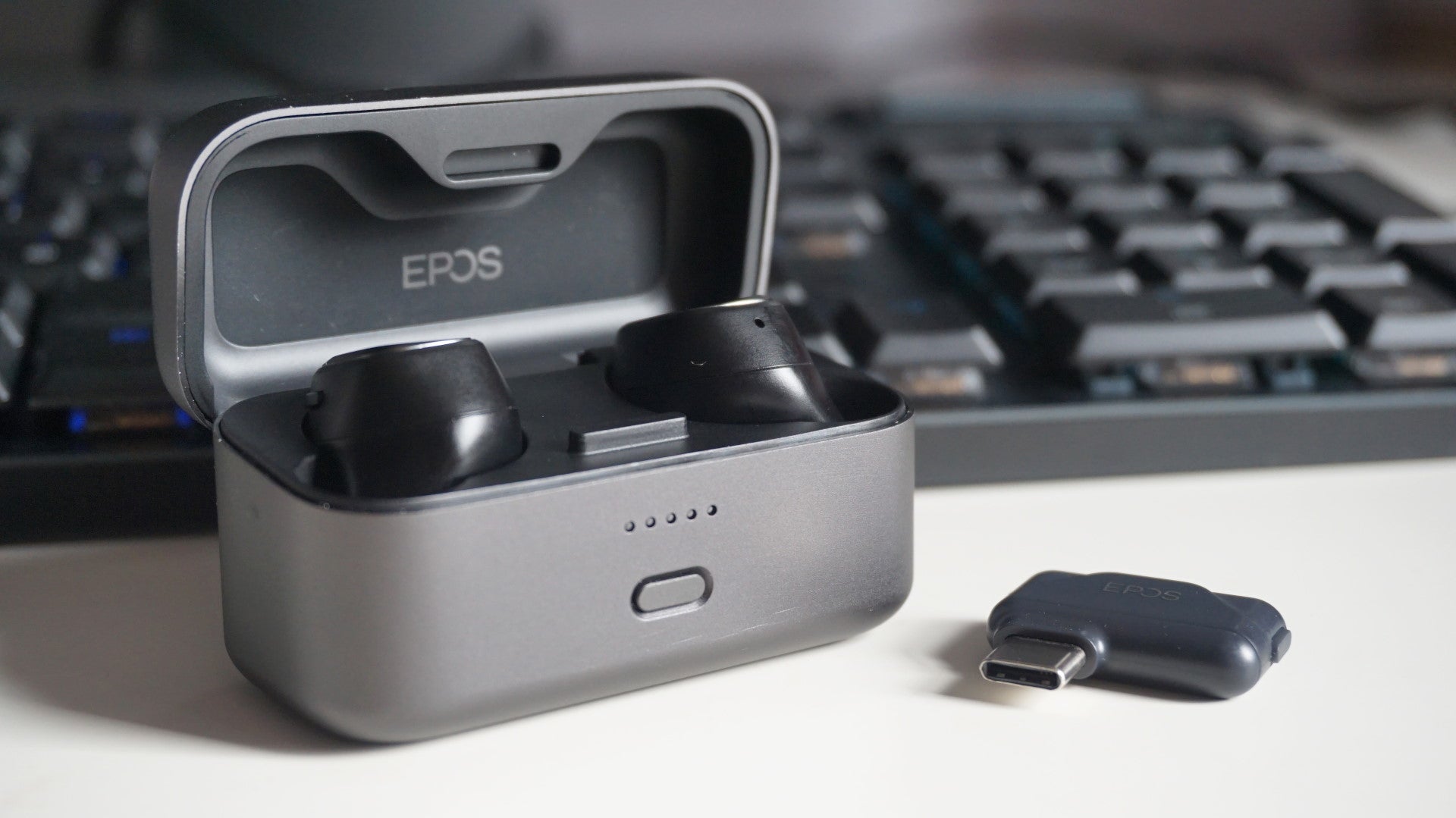 A photo of the EPOS GTW 270 Hybrid wireless earbuds inside their charging case, next to their USB-C adaptor