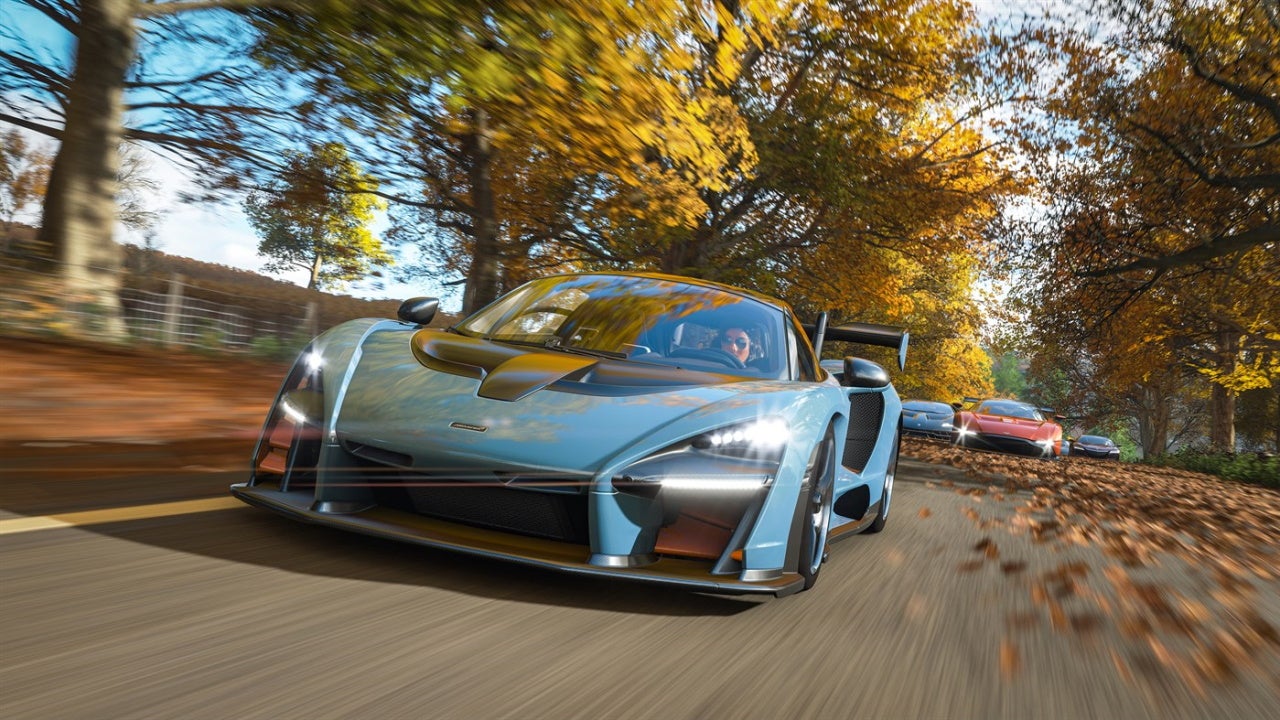 An image from Forza Horizon 4 which shows a blue supercar racing through a road surrounded by yellow autumn trees, and a cluster of other race cars lagging behind it.