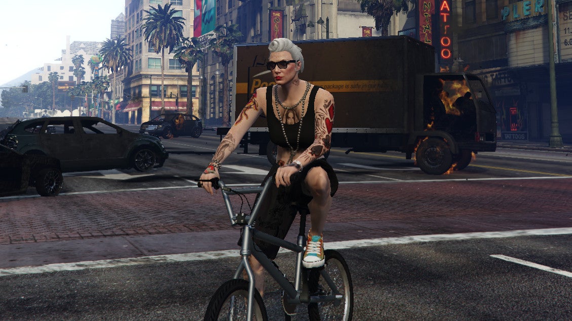 A woman sits on a BMX in front of burning vehicles in a GTA Online screenshot.