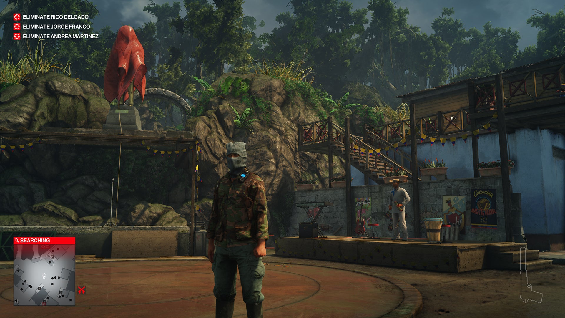 Ian Hitman in the Santa Fortuna level of Hitman 2. He is wearing camouflage combats and a balaclava, and is in a small town square in front of a stage, surrounded by jungle plants.