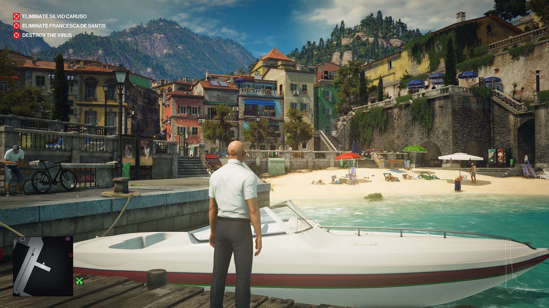A screenshot showing the Sapienza level of Hitman 2. Ian Hitman is standing on a jetty by a speedboat, looking at a lovely little beach and a quiet town in the sunshine.