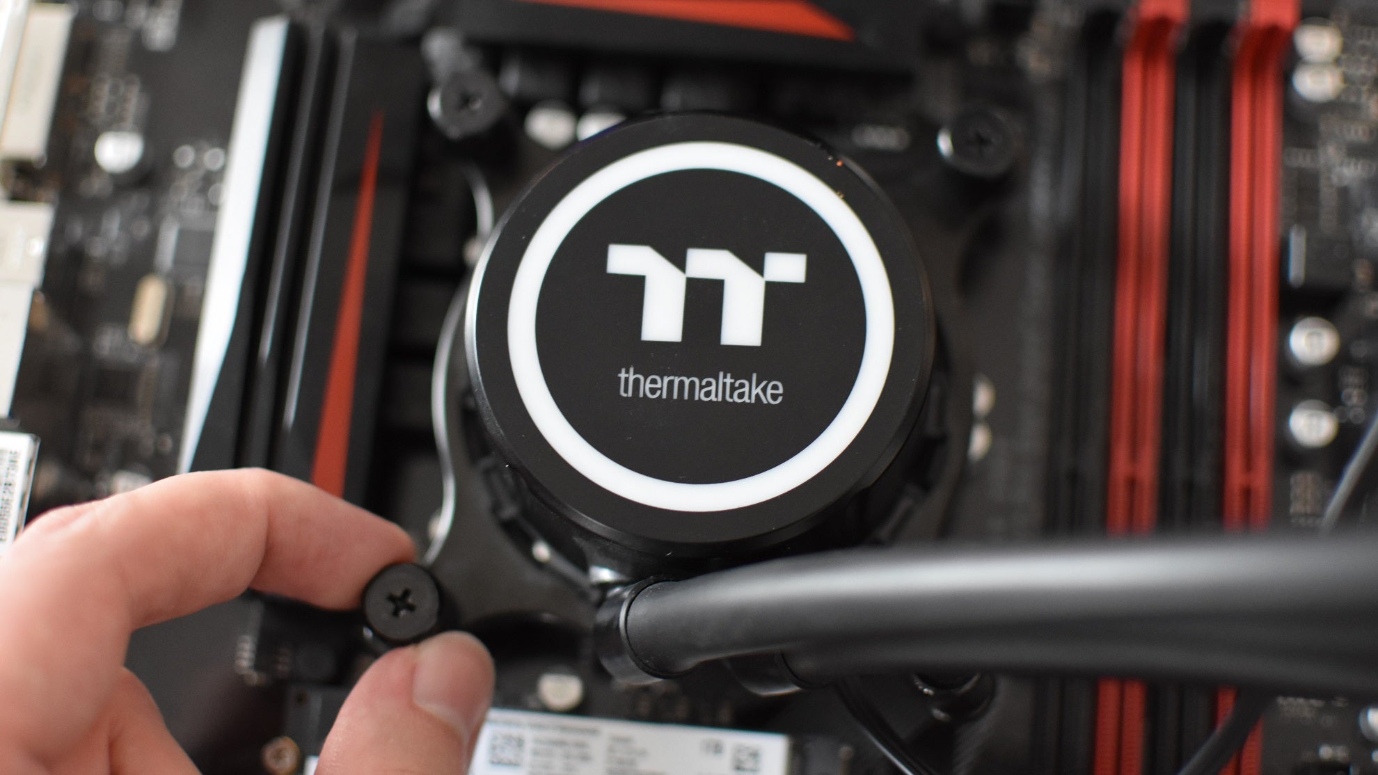 A hand tightens a thumbscrew on the mounting mechanism of a Thermaltake AIO watercooler.