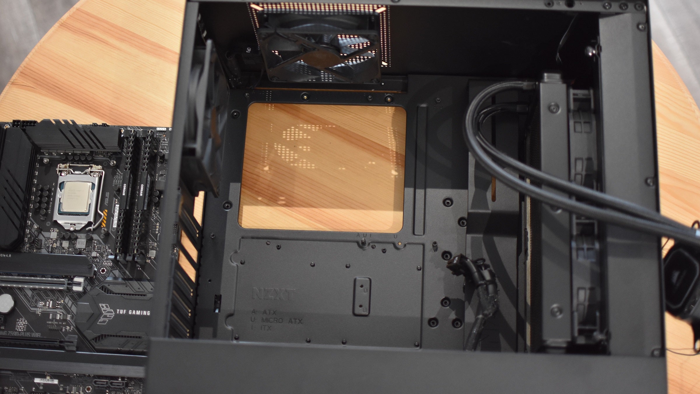 An empty PC case, showing the main internal chamber and motherboard tray. A motherboard sits next to the case on a table.