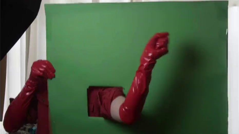 A hand in a red PVC glove waves out from a hole cut in a green screen.