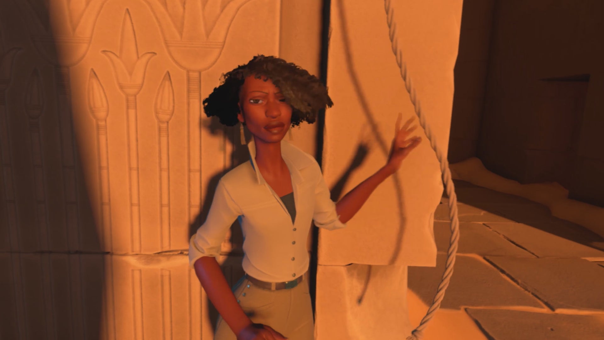 A screen from In The Valley Of Gods showing a black woman with a short bob and wearing a khaki shirt and trousers, standing at golden hour in front of part of a stone ruin
