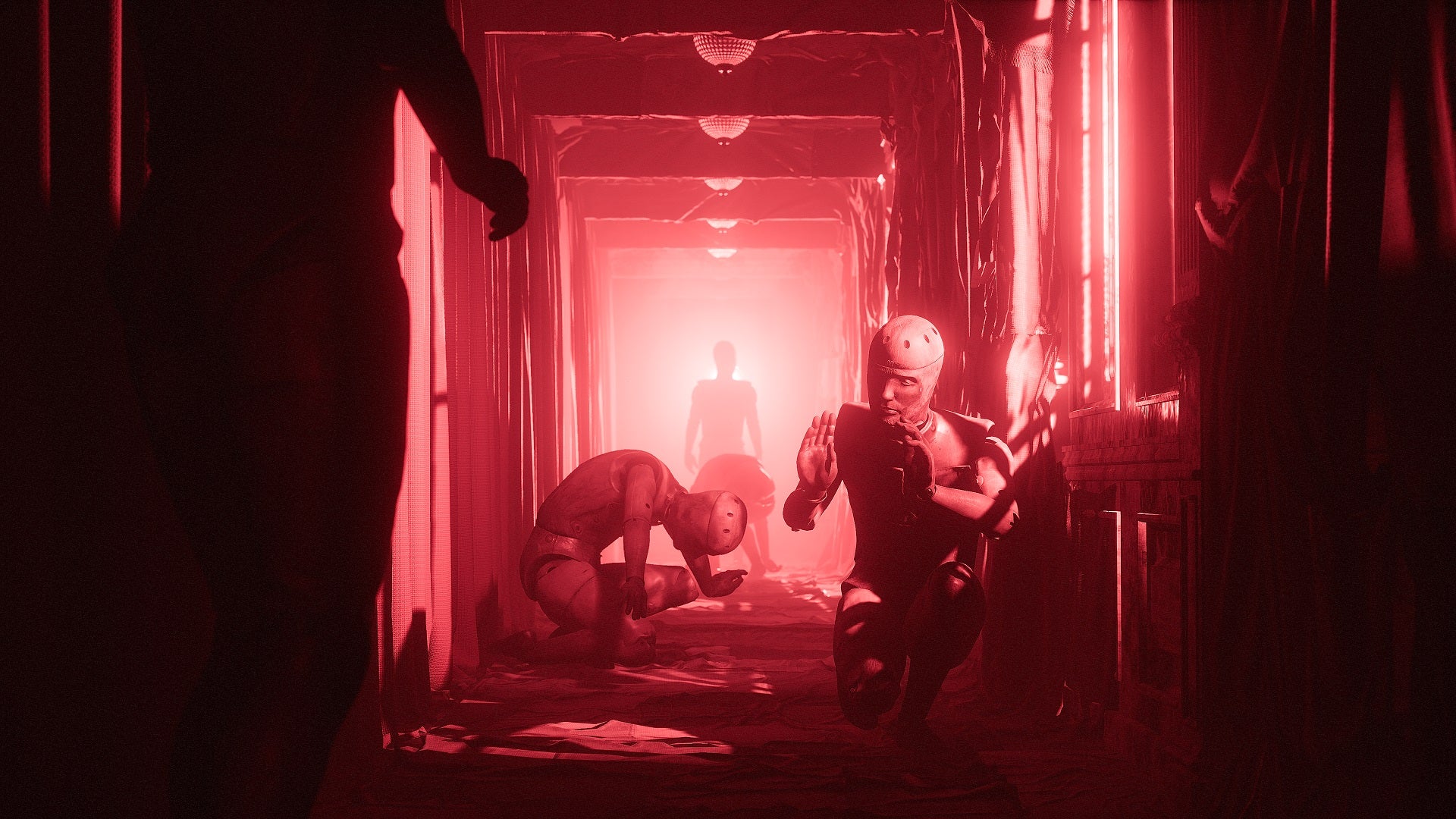 In an entirely red-lit corridor, several mannequins cower in poses of fear and submission. Two more humanoid figures can be seen standing at either end of the corridor in more confident poses, although it's hard to say if they're mannequin or human.