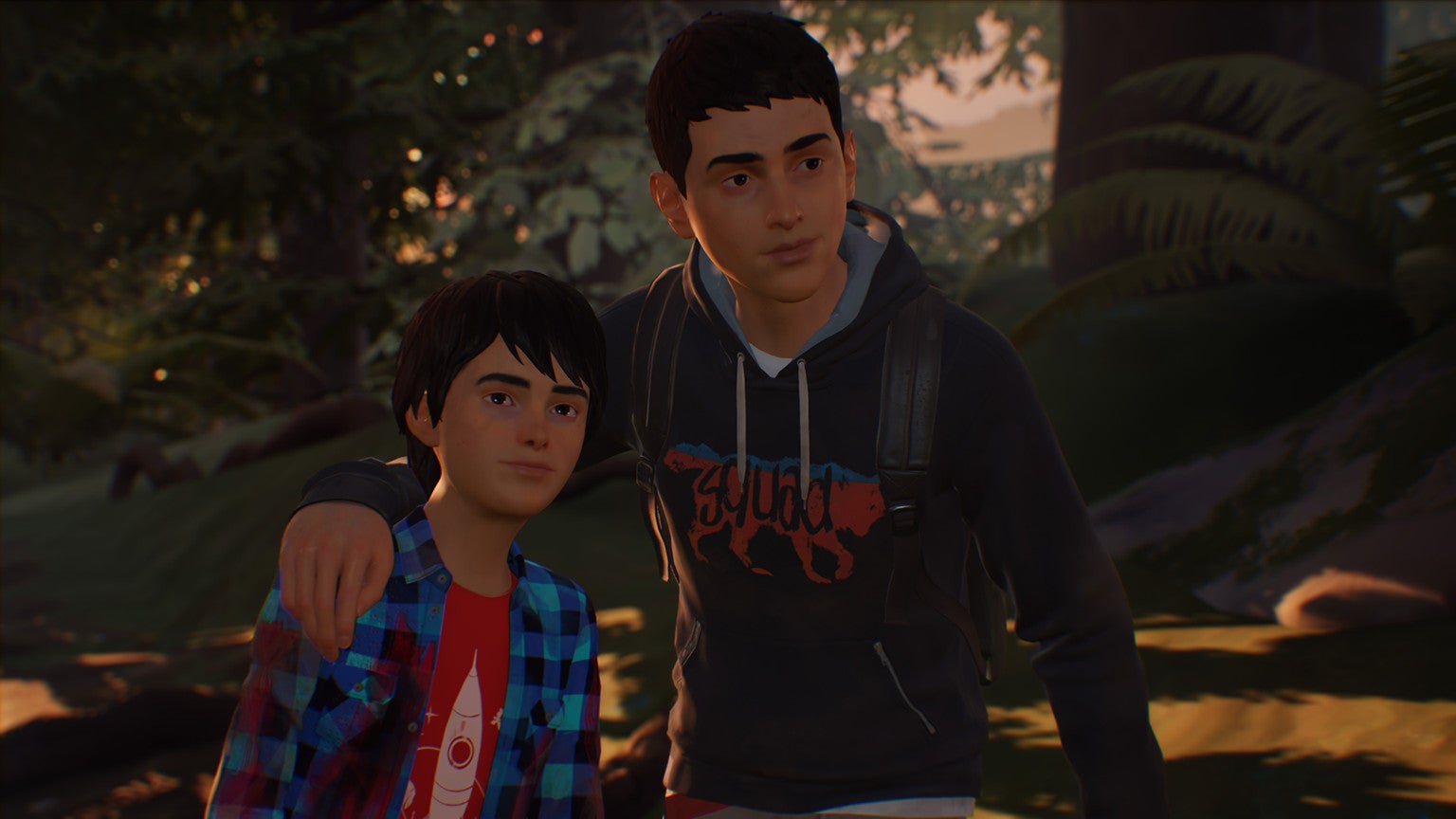 The protagonists of Life Is Strange 2 in a close-up against a forest background. Older brother Sean has his arm around younger brother Daniel, and both are smiling slightly.
