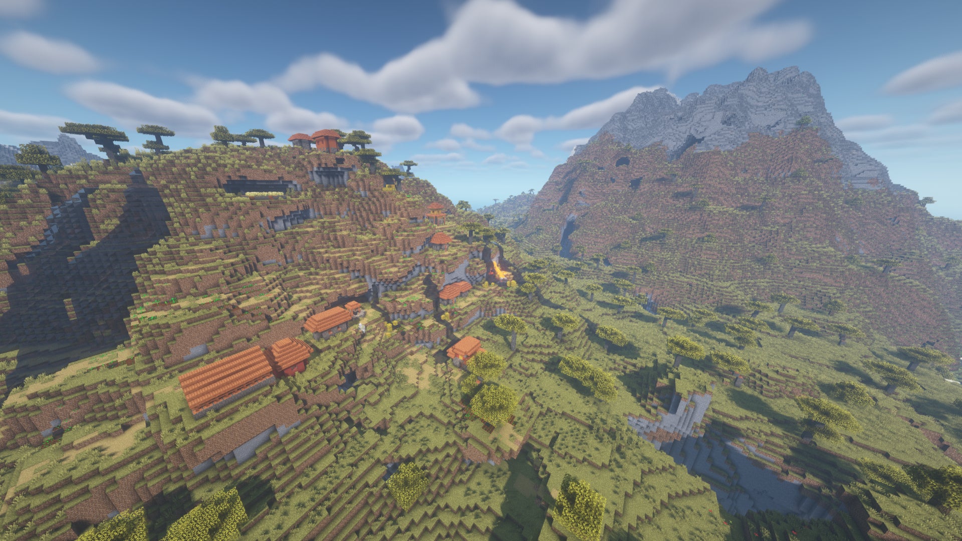 A Minecraft village built across the side of a savanna hill, with a large stone-topped mountain in the background.
