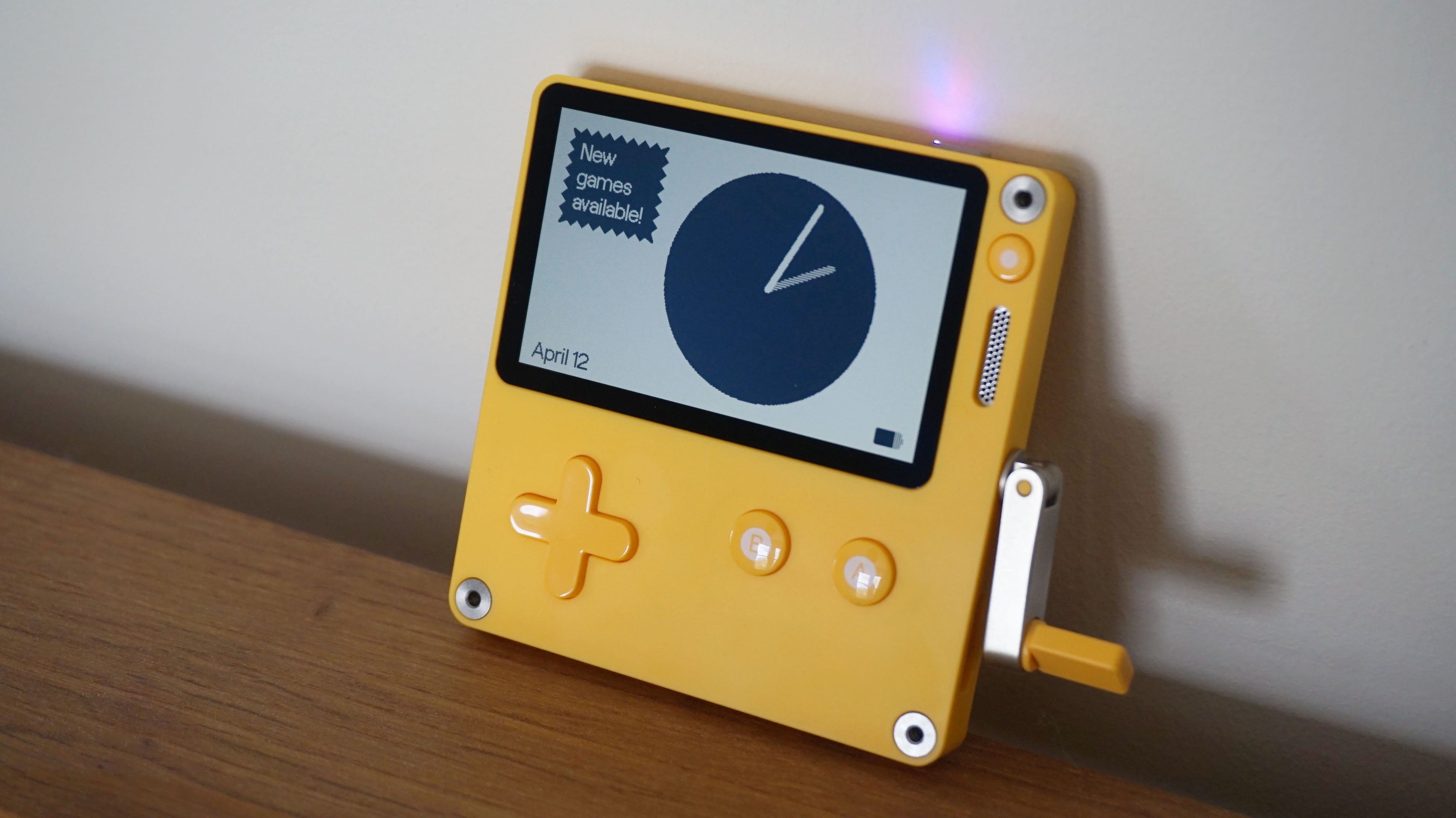 The Playdate leans against a white wall, showing the standby clock onscreen and a banner saying new games are available