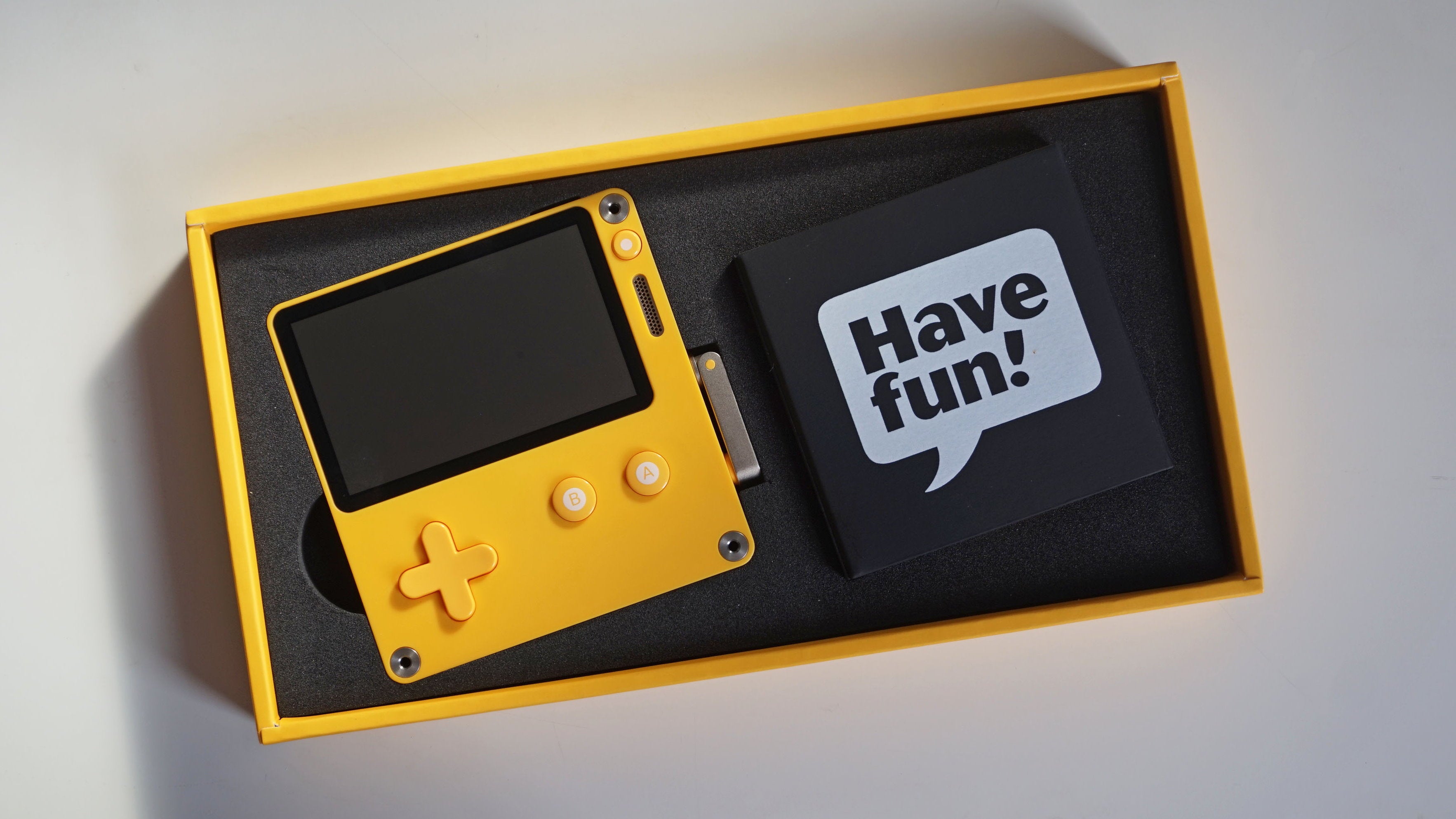 The Playdate inside its consumer packaging, with a cardboard USB wrapper saying 'Have Fun!' next to it