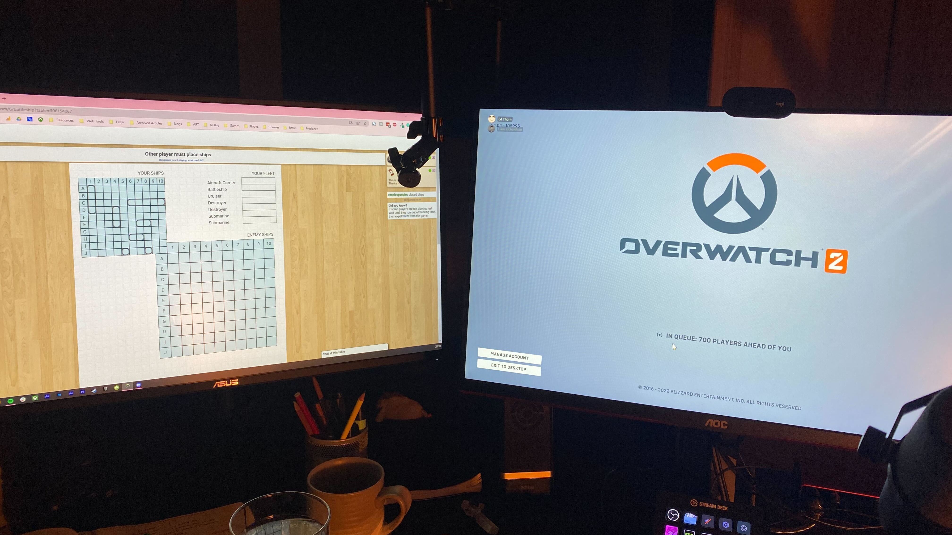 A photo of a desk with two PC monitors: one on the left queuing for Overwatch 2, the other playing a game of Battleship