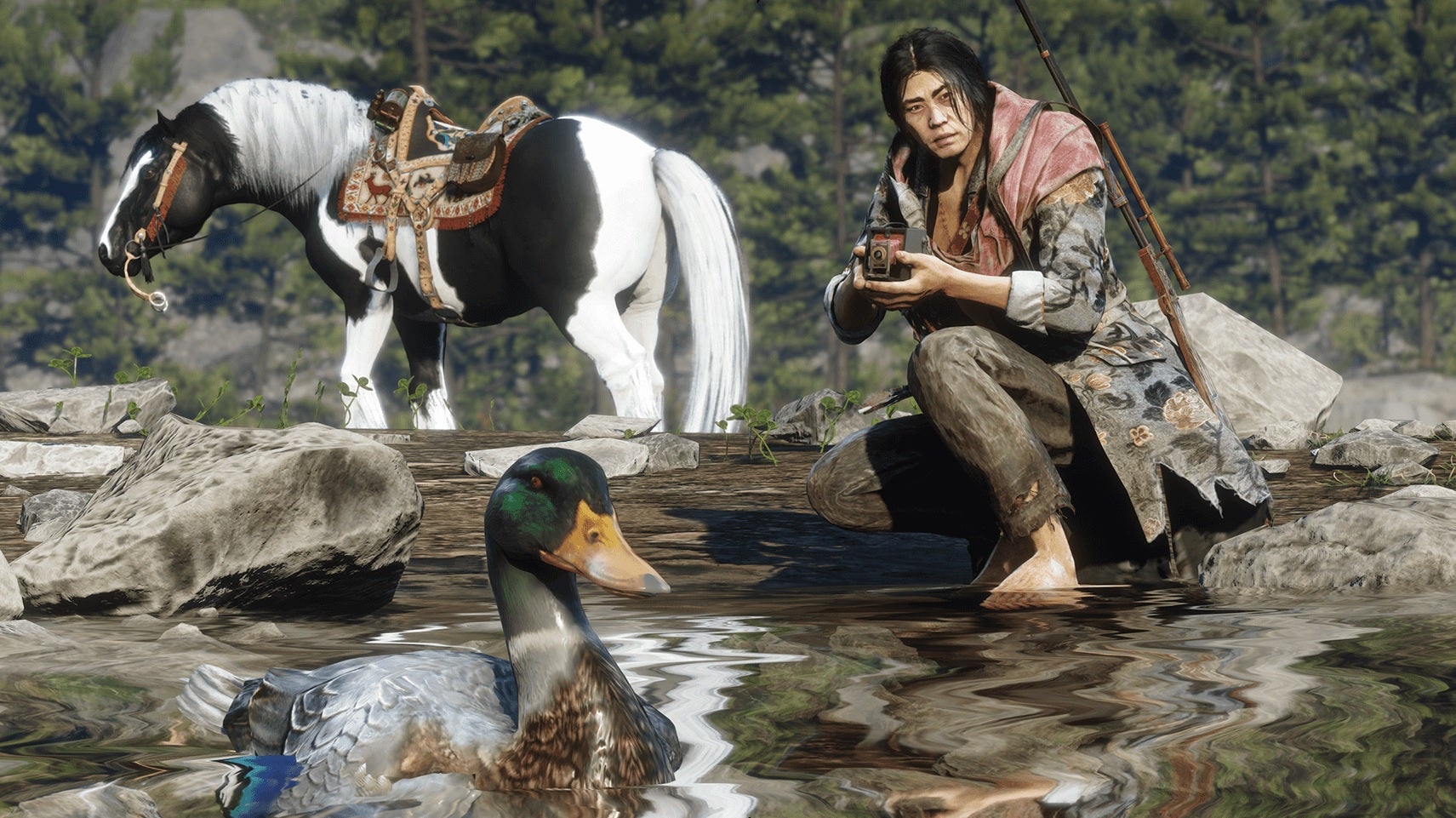 Red Dead Redemption 2 Online - A naturalist player crouches near the water holding a camera with a duck in the foreground while their horse waits in the background.