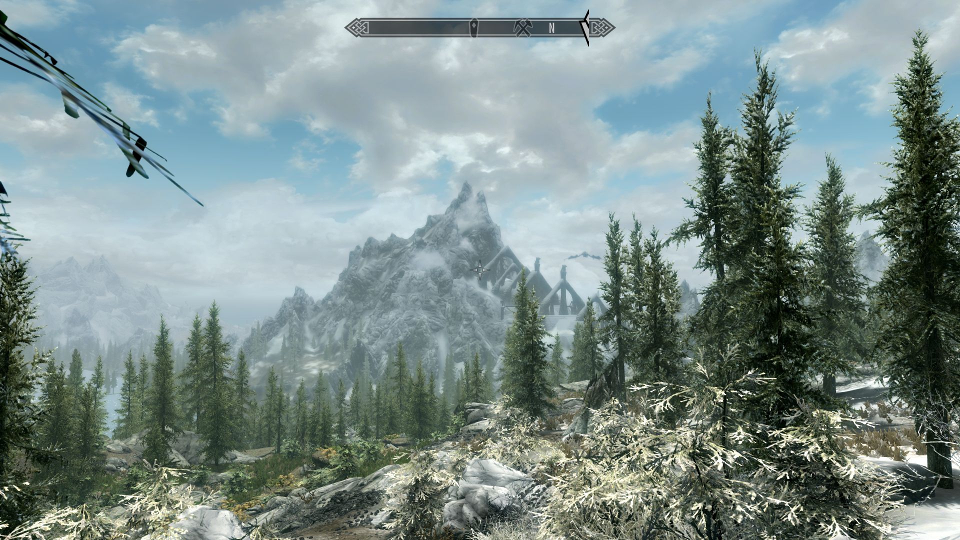 Eine Ansicht in The Elder Scrolls V: Skyrim mit einem blauen Wolkenhimmel, einem Kiefernwald im Vordergrund und einem schneebedeckten Berg in der Ferne