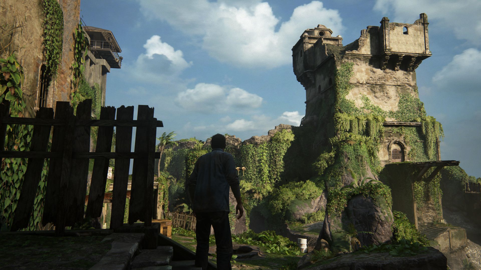 Protagonist Nate Drake looking at a large and ruined prison tower, covered in plants, off in the distance, in an early level of Uncharted 4: A Thief's End
