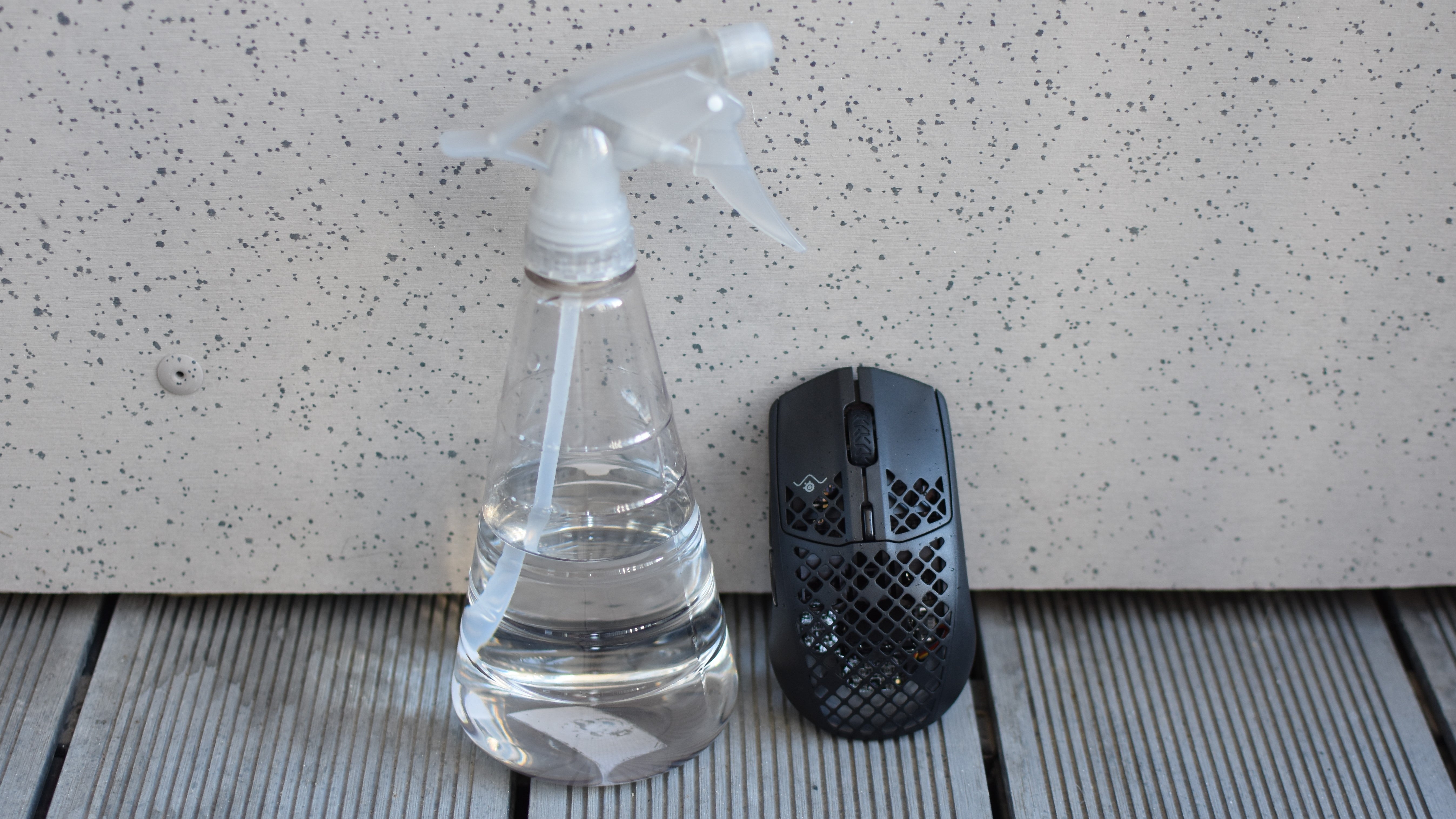 A partially wet SteelSeries Aerox 3 Wireless mouse sitting upright next to a spray bottle of water.